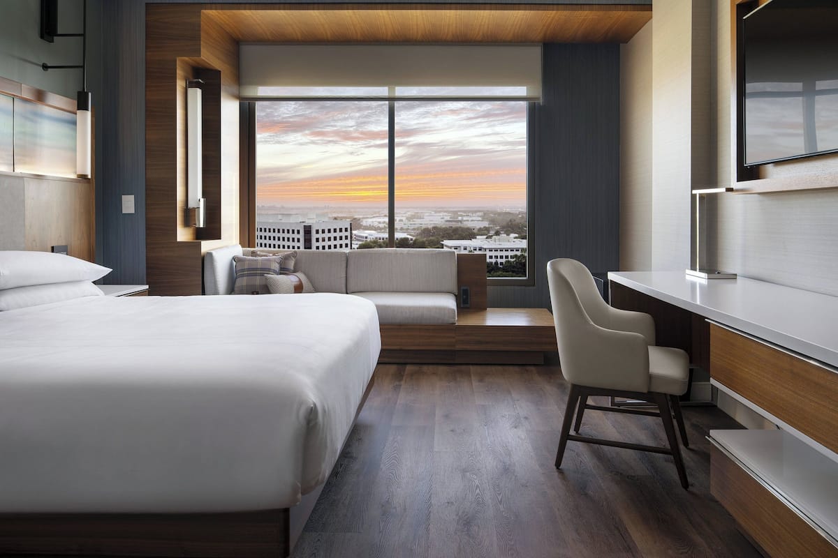 A modern hotel room with a large bed, a desk, and a view of the city at sunset.