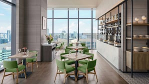 Un espace repas moderne doté de plusieurs tables et chaises, d’un comptoir de cuisine avec des ustensiles, et de grandes fenêtres offrant une vue sur la ville.