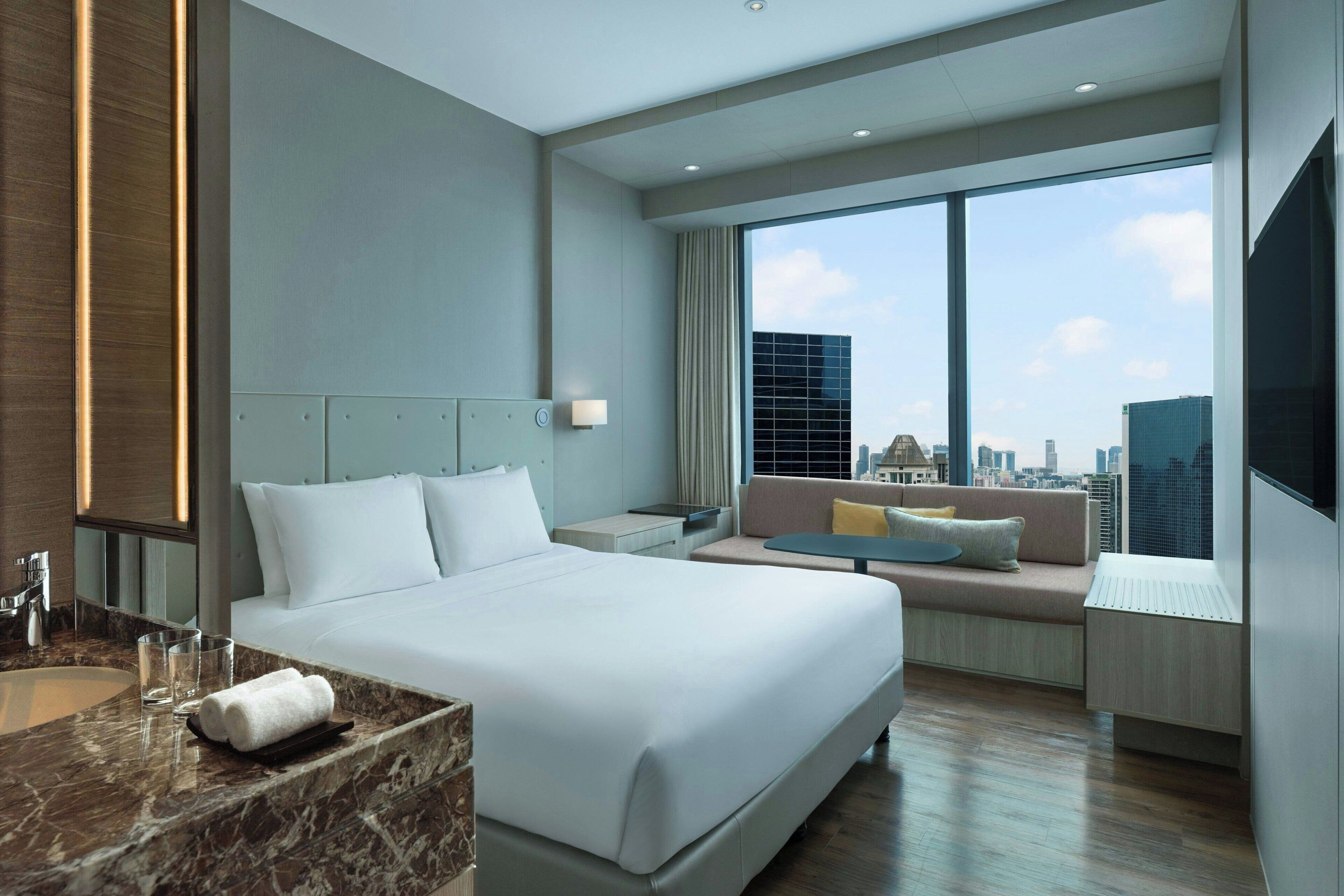 A modern hotel room with a large bed, a seating area with a sofa and armchairs, a large window with a city view, and a marble bathroom counter.