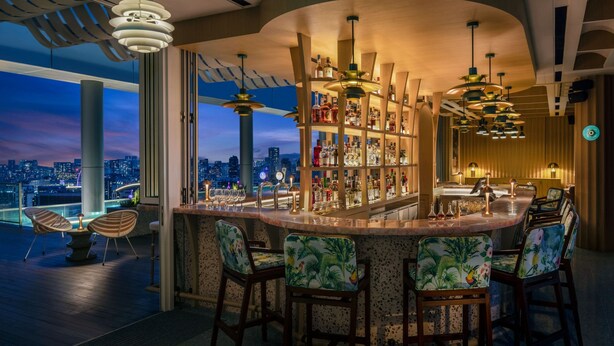 Un espace bar offrant une vue sur la ville, doté d’un éclairage moderne et de chaises à thème tropical.