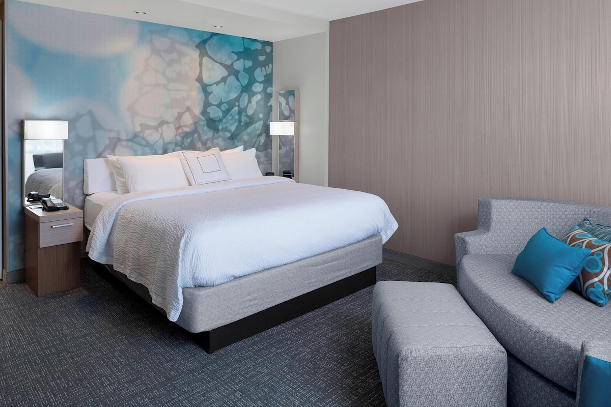 A modern hotel room with a large bed, a sofa, and a nightstand with a lamp.
