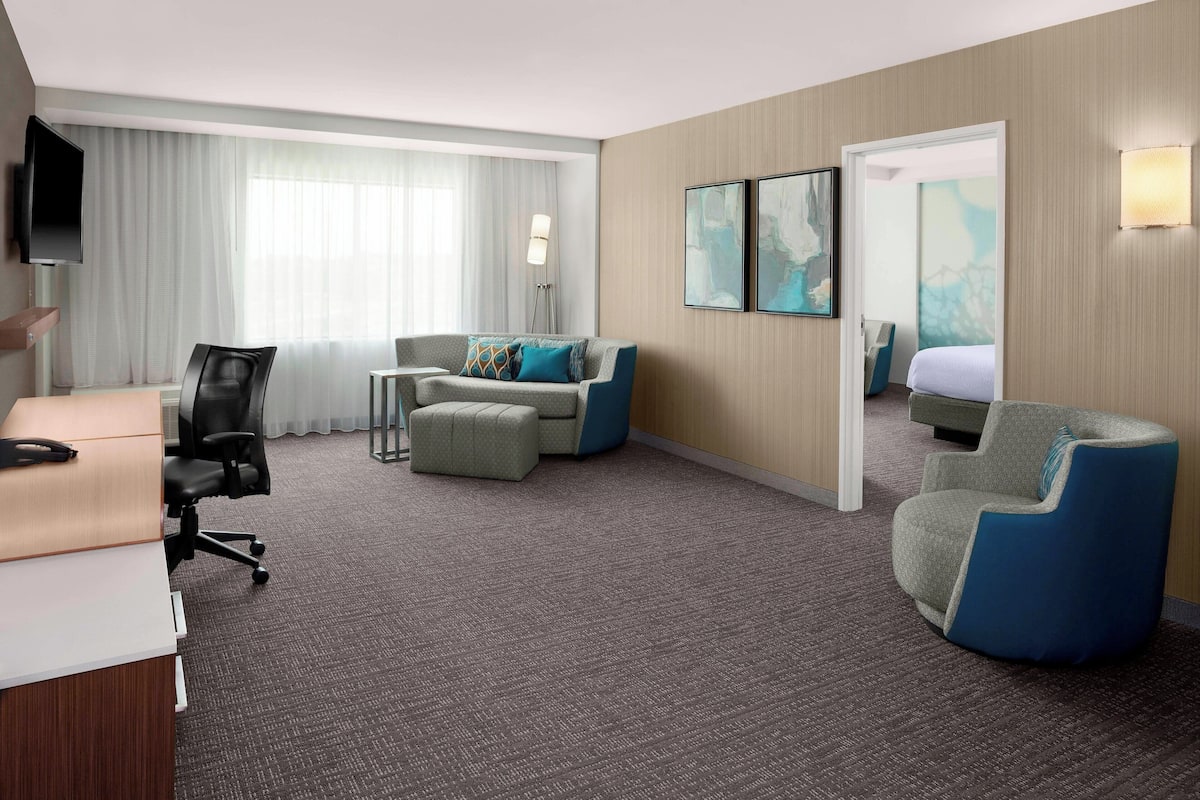 A hotel room with a desk, two chairs, a sofa, a bed, a TV, and artwork on the walls.