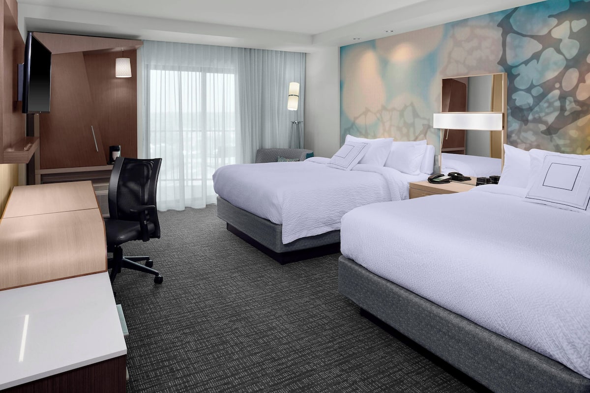 A hotel room with two beds, a desk, a chair, a TV, and a large window with curtains.