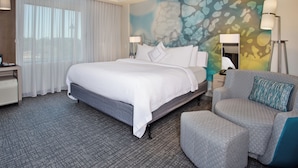 A hotel room with a large bed, a grey armchair, a desk with a lamp, and a wall with a textured mural. - Courtyard by Marriott Dallas Flower Mound (Flower Mound)