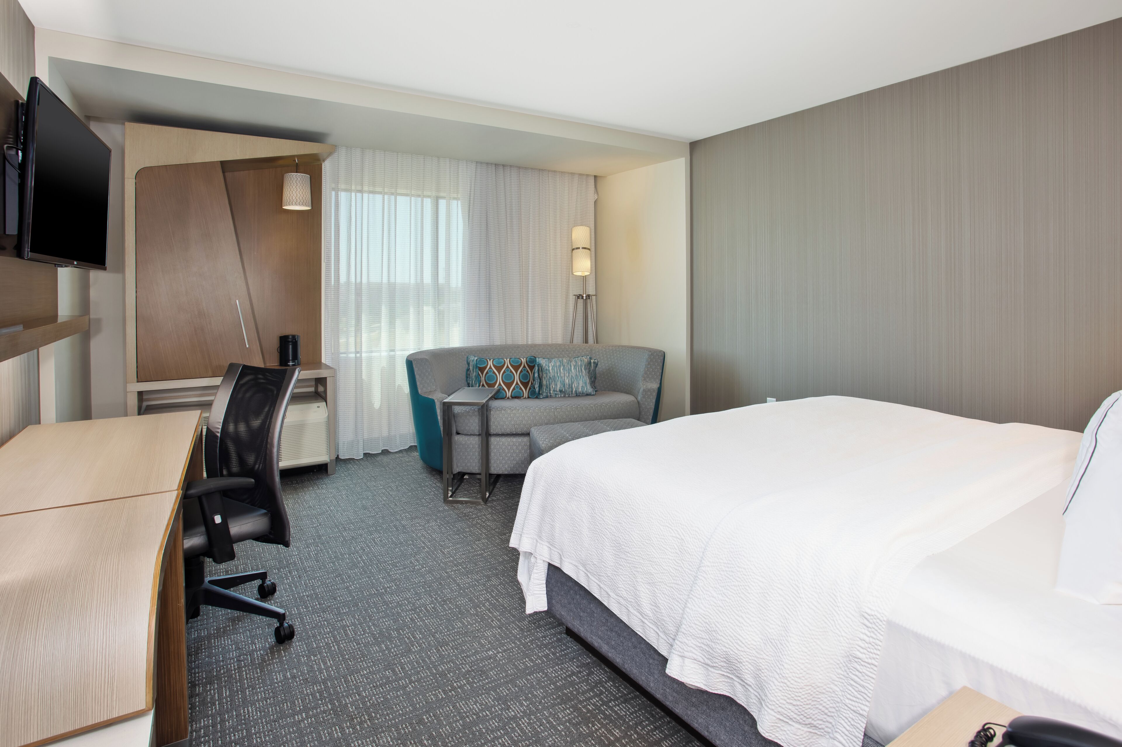 Une chambre d’hôtel comprenant un lit, un bureau avec une chaise, un canapé, un téléviseur fixé au mur et une lampe.