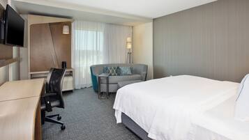 Une chambre d’hôtel comprenant un lit, un bureau avec une chaise, un canapé, un téléviseur fixé au mur et une lampe.