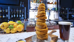 Un montón de aros de masa frita sobre una tabla de madera, un vaso de cerveza y una cesta de limones y limas en una encimera de mármol.