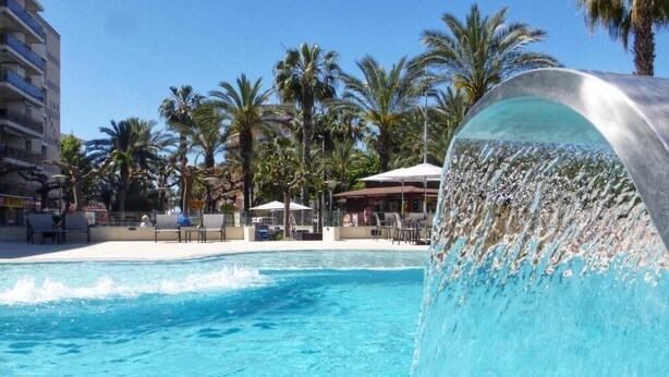 A swimming pool with a waterfall feature, surrounded by palm trees and a building with a white facade. - Rentalmar Navarra (Salou)