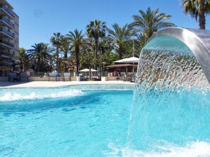Una piscina con cascada, rodeada de palmeras y un edificio de fachada blanca.