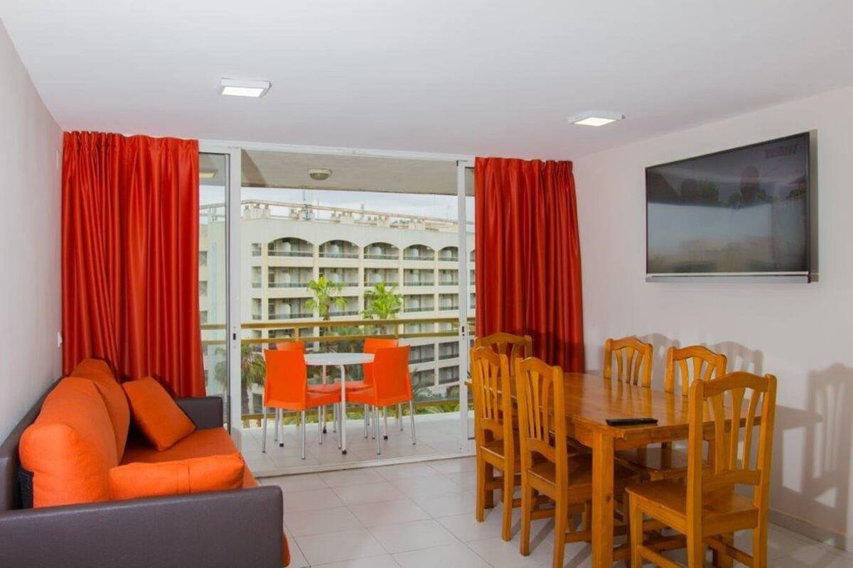 A room with a sofa, dining table, chairs, and a balcony with orange chairs.