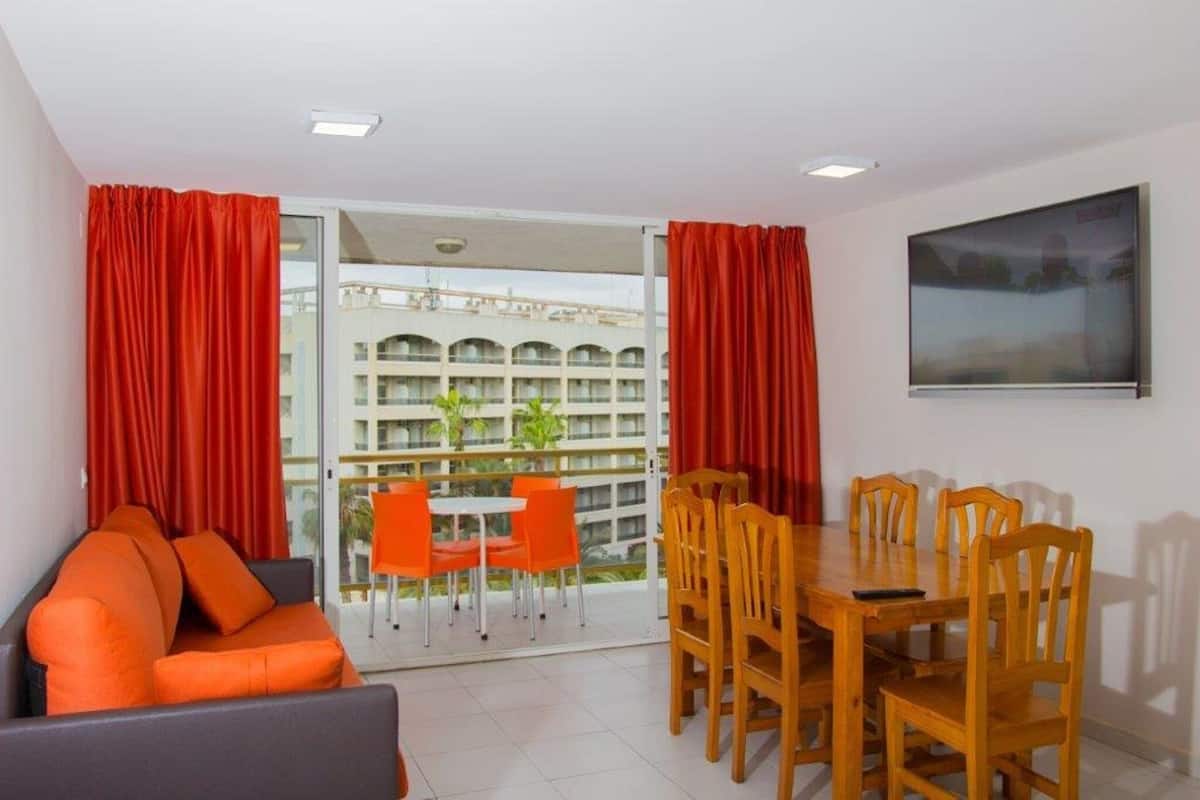 A room with a sofa, dining table, chairs, and a balcony with orange chairs.