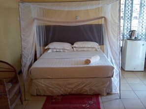 A bed with a canopy, pillows, a rolled towel, a small refrigerator, and a chair. - Zambezi Way Lodge (Livingstone)