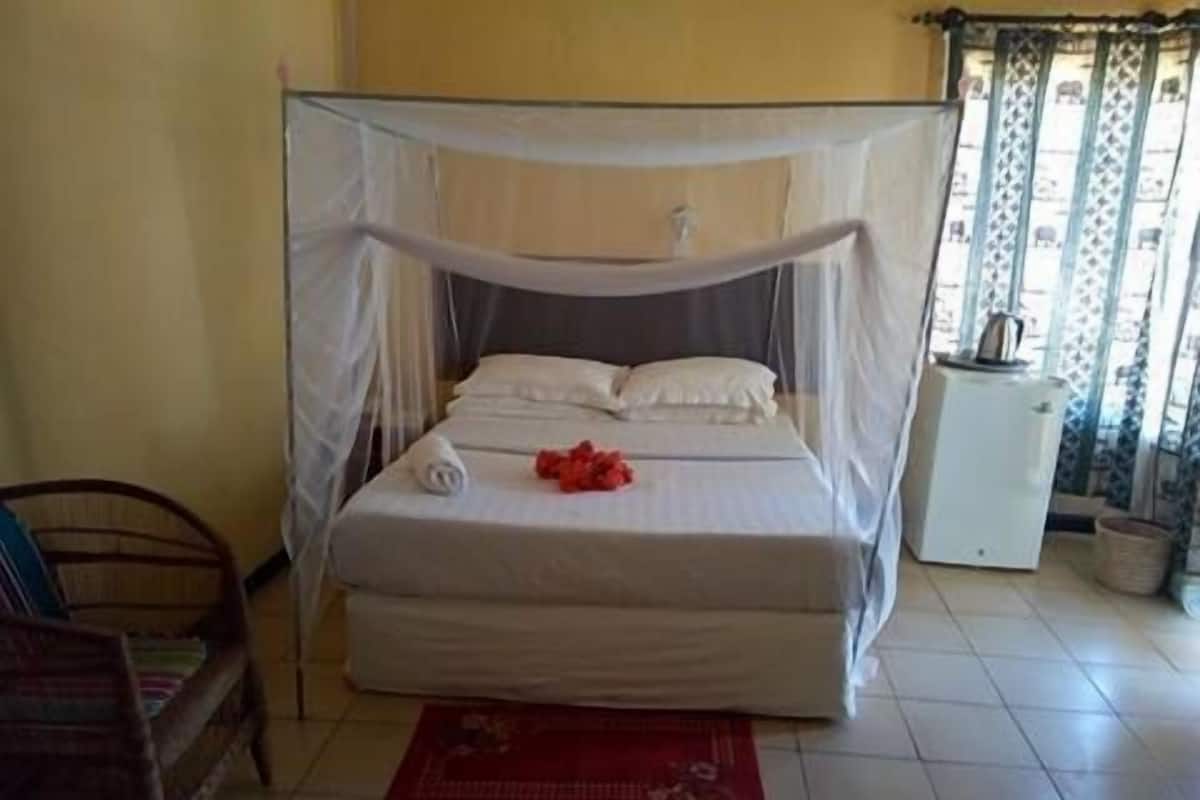 A single bed with a mosquito net, white bedding, and a red mat in a room with tiled flooring.