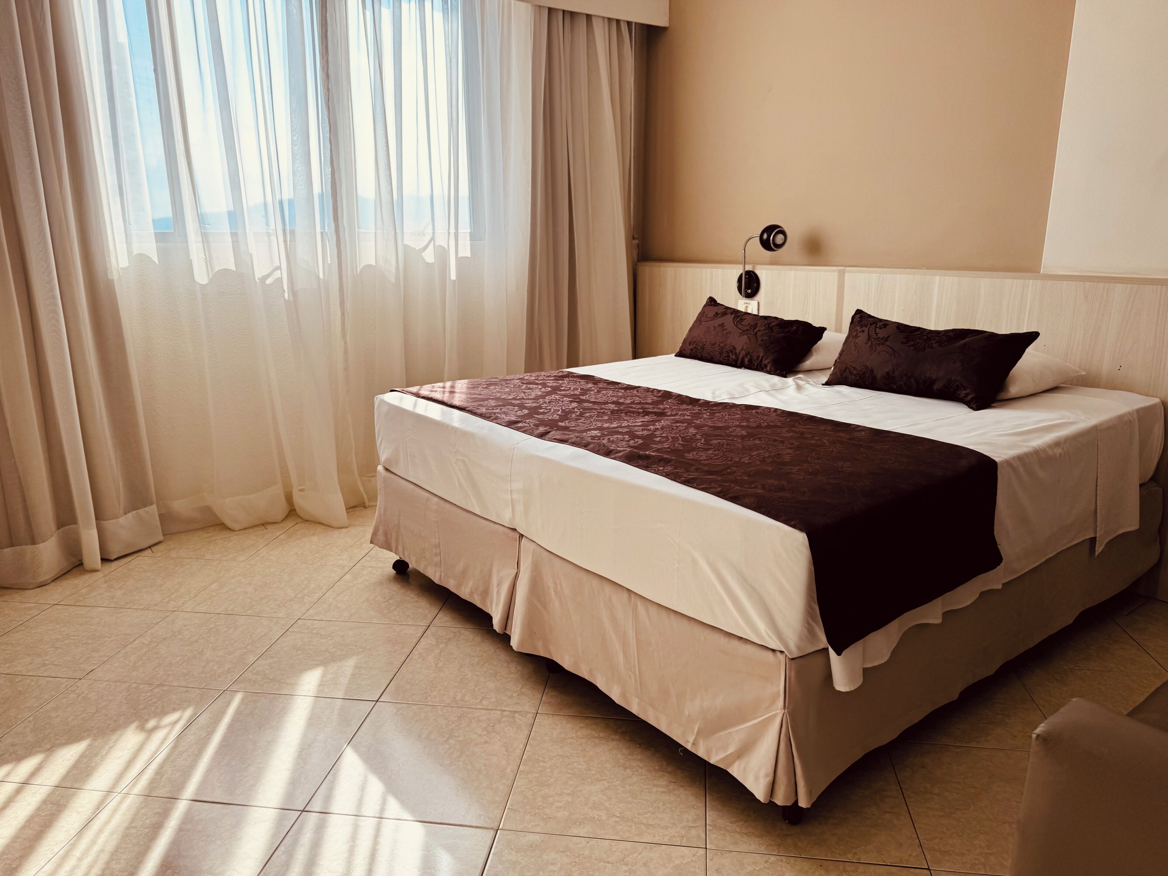 Habitación de hotel con una cama grande, cortinas, una mesita de noche y una lámpara de pared.
