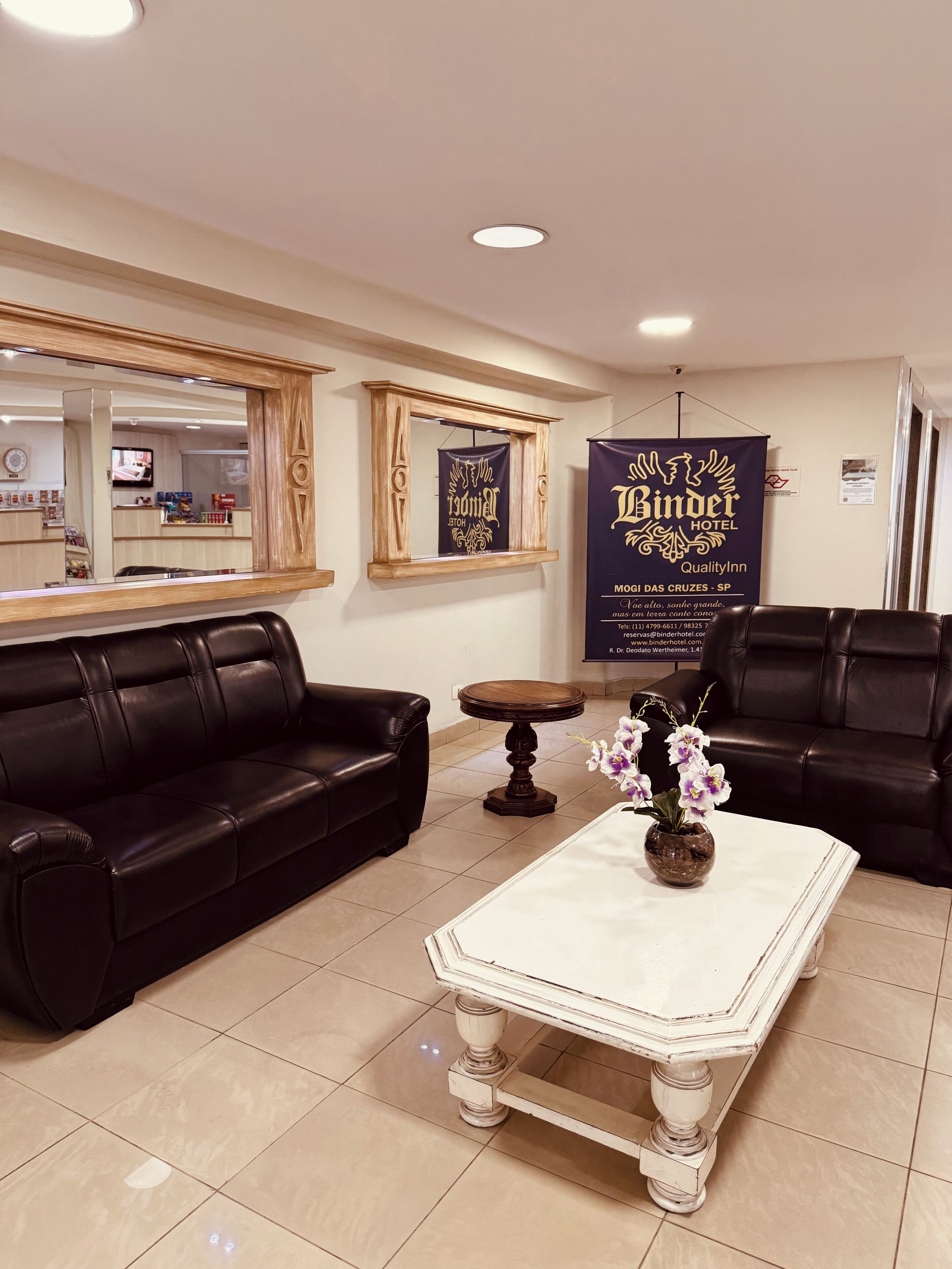A hotel lobby with two black leather sofas, a white coffee table, a wooden side table, and a reception desk with a mirror.