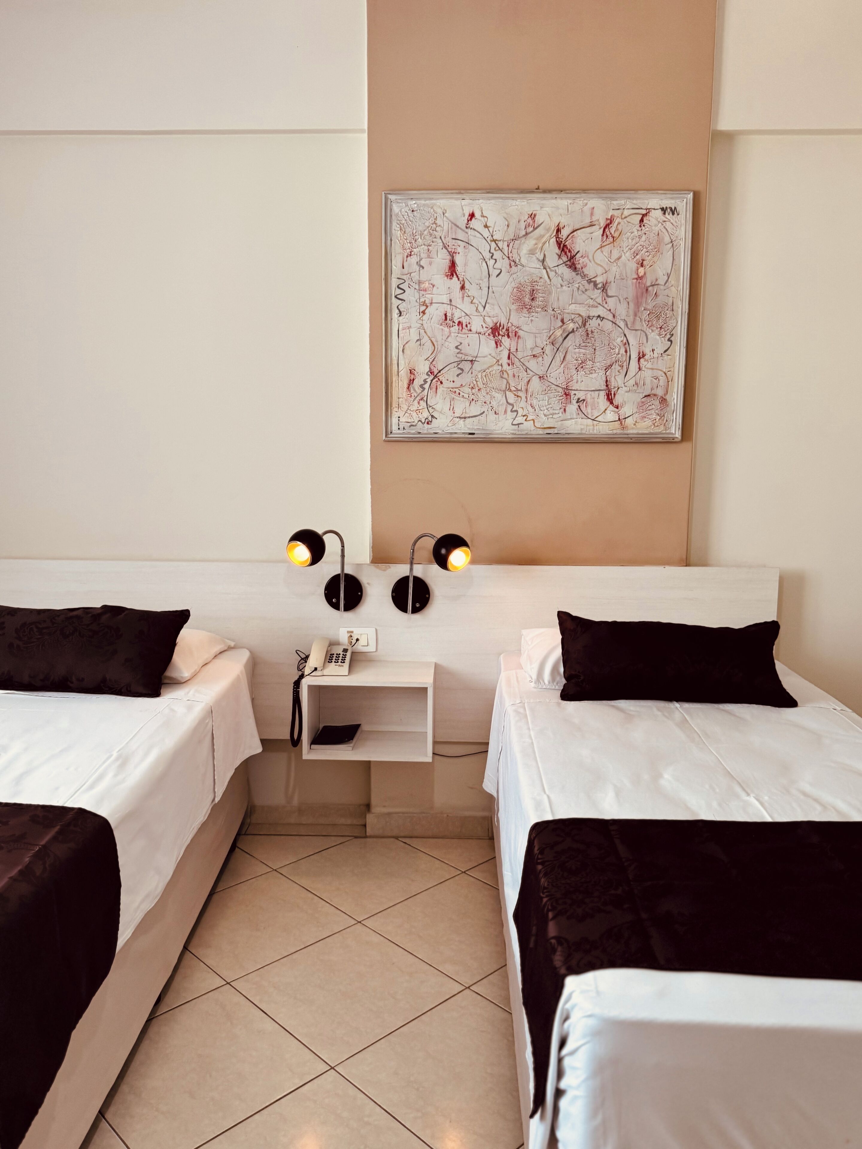 A hotel room with two beds, a nightstand with a phone and lamp, and a wall art piece.