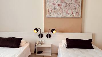 Quarto de hotel com duas camas, uma mesa de cabeceira com telefone e luminária, e uma obra de arte na parede.