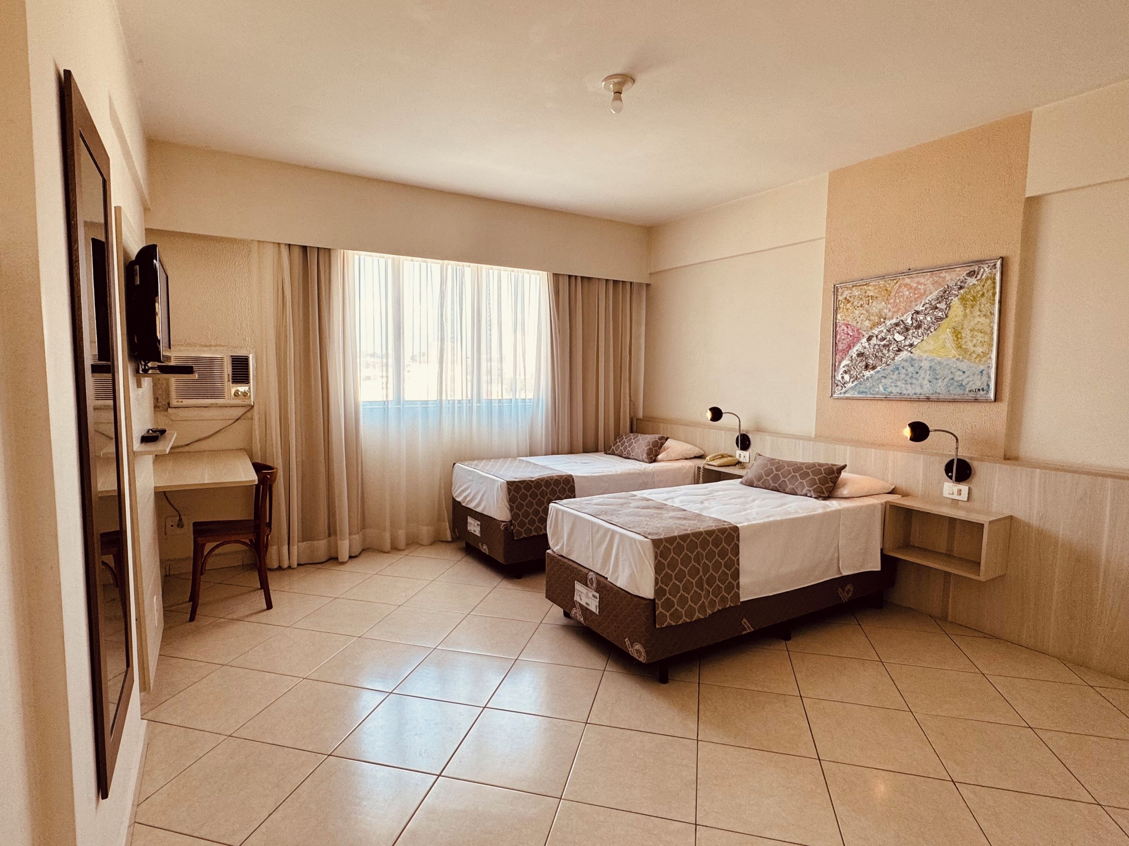 A hotel room with two beds, a desk, a chair, and a large window with curtains.