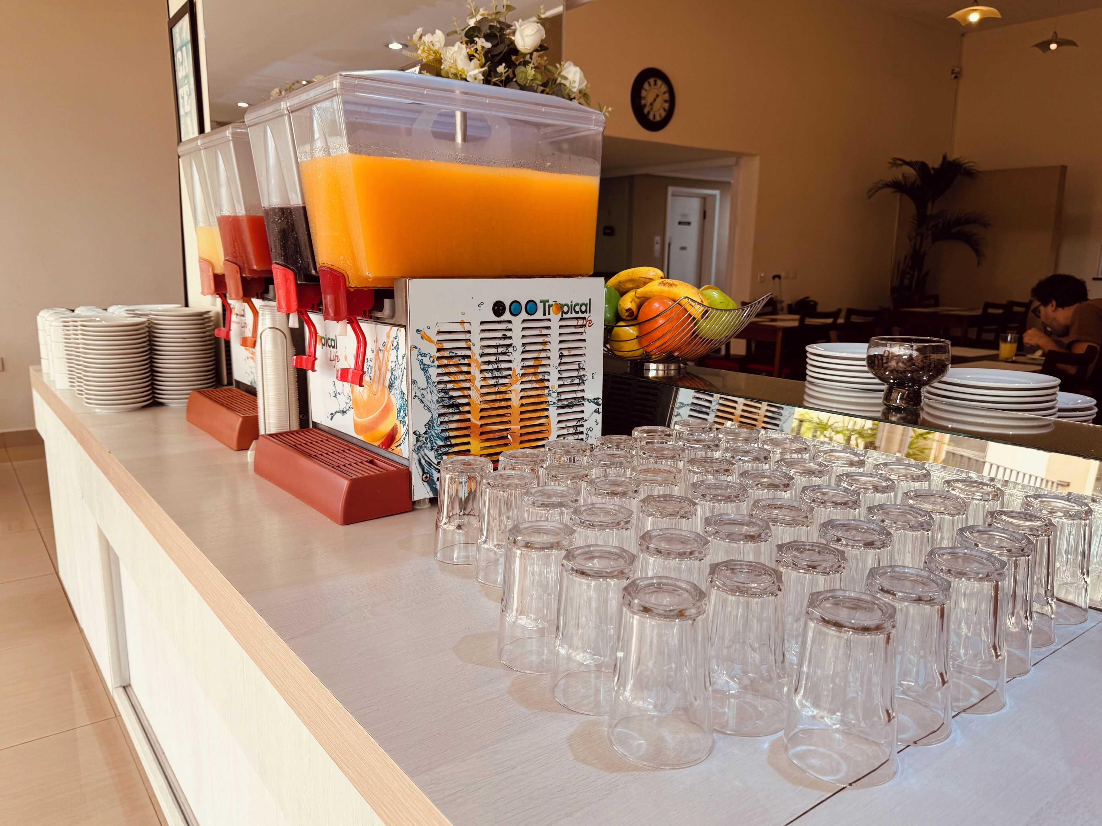 A buffet area with a juice dispenser, stacked plates, and a fruit bowl.