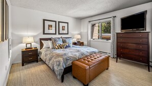 A bedroom with a bed, two bedside tables with lamps, a dresser, a television, and a window with curtains.