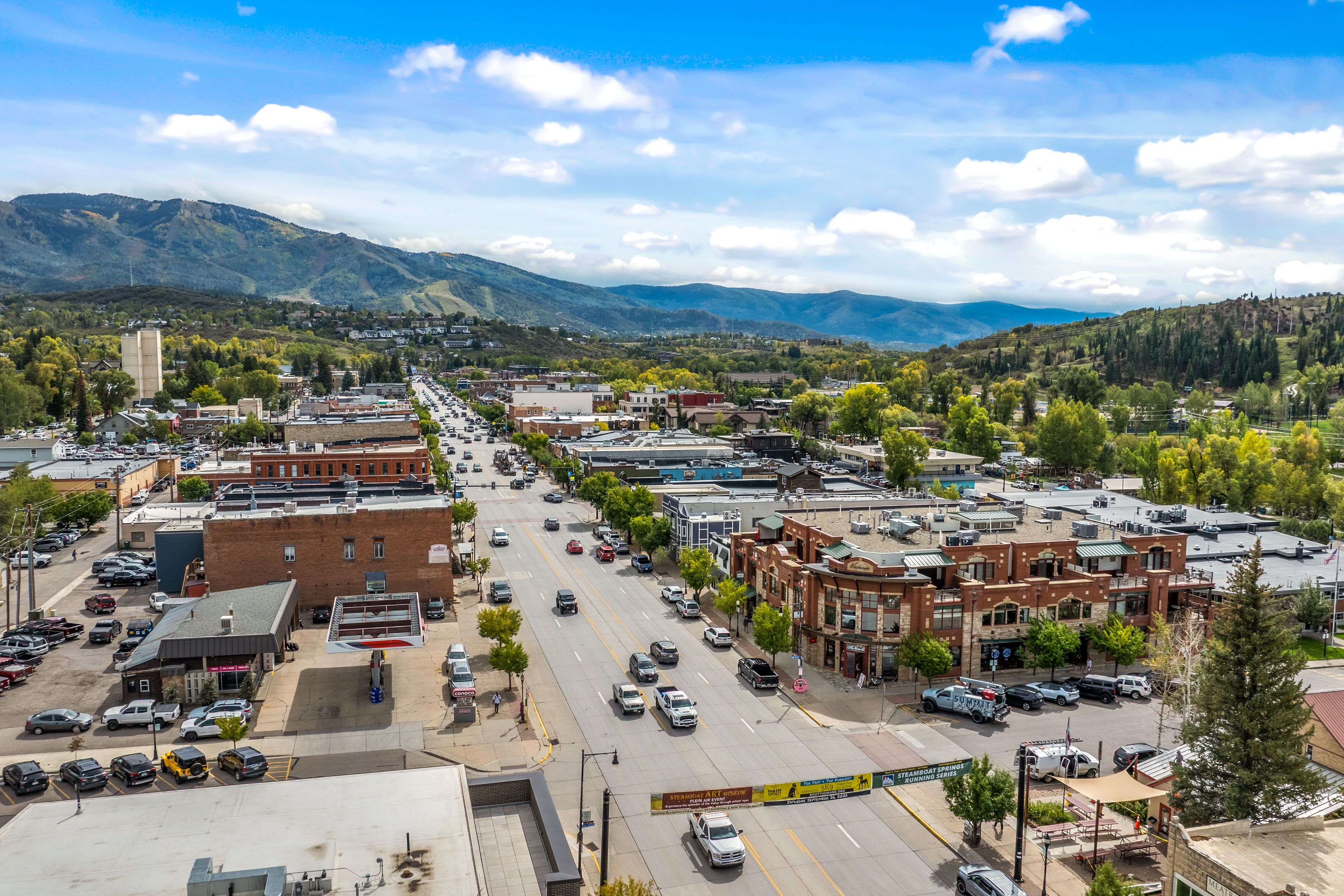 A town with a main street lined with buildings, a parking lot, and a mountainous backdrop.