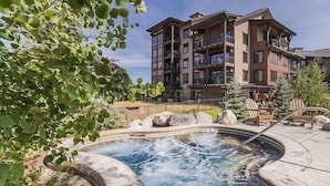 A hot tub surrounded by rocks and trees, with a multi-story building in the background. - First Tracks 3301 2 Bedroom Condo by RedAwning (Steamboat Springs)