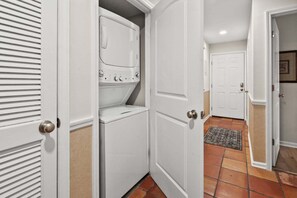 A laundry room with a stacked washer and dryer, a door with a round handle, and a tiled floor. - Sea Cloisters 101F by RedAwning (North Myrtle Beach)