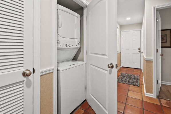A laundry room with a stacked washer and dryer, a door with a round handle, and a tiled floor. - Sea Cloisters 101F by RedAwning (North Myrtle Beach)