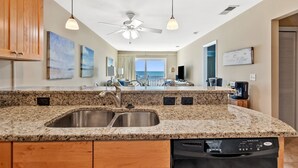 A kitchen with granite countertops, stainless steel sink, wooden cabinets, a ceiling fan, pendant lights, and a view of the ocean through a large window. - Sea Cloisters 304F by RedAwning (North Myrtle Beach)