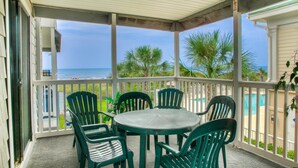 A covered outdoor area with a table and chairs, overlooking a pool and palm trees. - Sea Cloisters 201D by RedAwning (North Myrtle Beach)