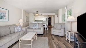 A living room with a sofa, armchairs, a coffee table, a TV, and a dining area with a kitchen in the background. - Shoreham Towers 5C by RedAwning (North Myrtle Beach)