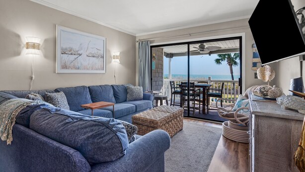 A modern living room with a blue sofa, a small table, and a view of the ocean through a sliding glass door. - Sea Cloisters 201E by RedAwning (North Myrtle Beach)