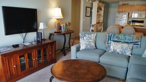 A living room with a television, a wooden cabinet, a coffee table, and a sofa with patterned cushions. - Sea Cloisters 305C by RedAwning (North Myrtle Beach)