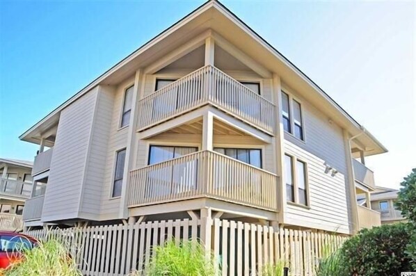 A multi-story residential building with a corner balcony, a picket fence, and a parked car. - Tilghman Lakes B3 by RedAwning (North Myrtle Beach)