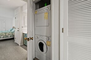 A laundry room with stacked washers and dryers, a bed with patterned bedding, and a door with a brass doorknob. - Tilghman Lakes B3 by RedAwning (North Myrtle Beach)