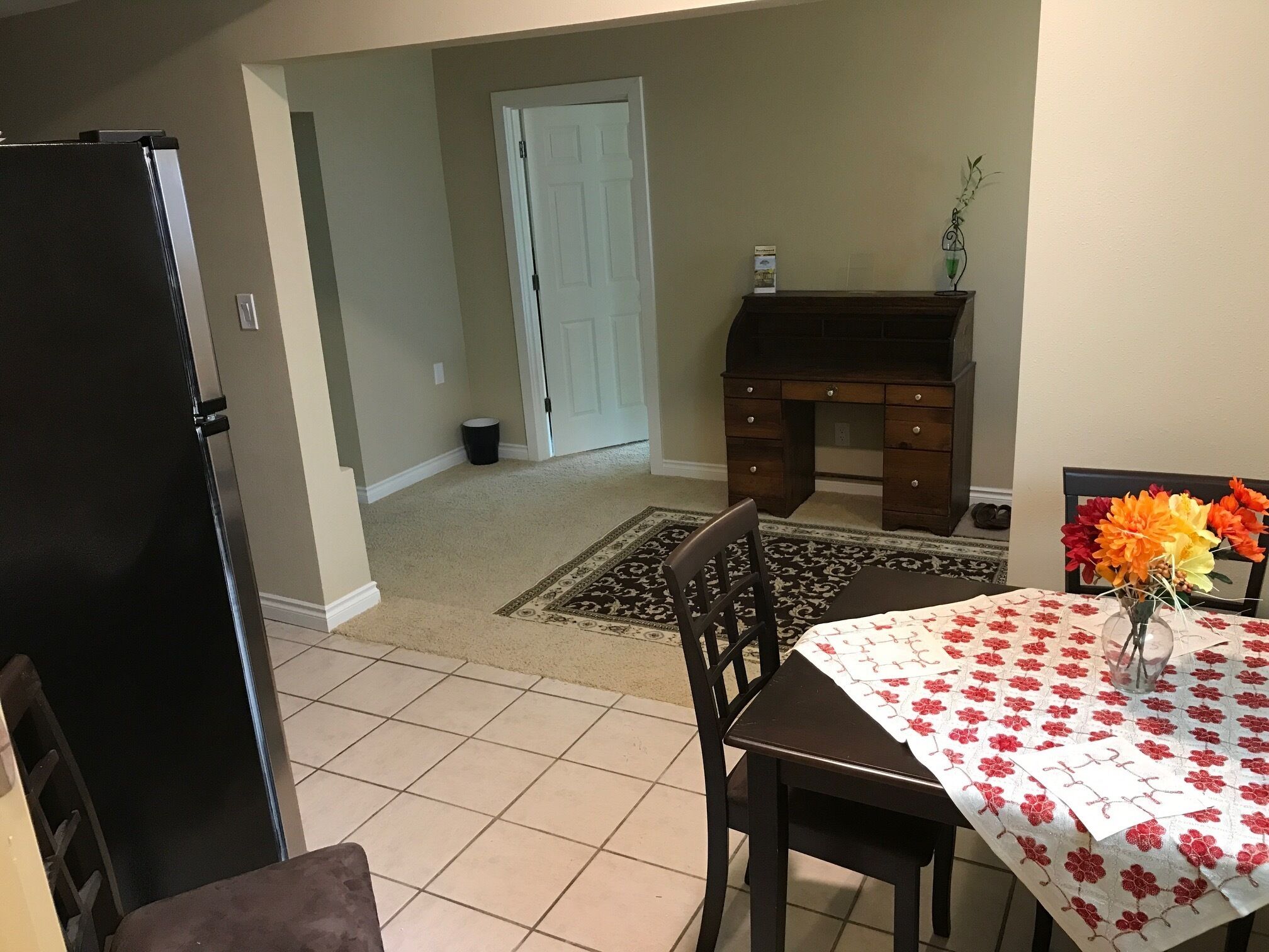 A dining area with a table set for two, a dark wooden desk, and a black refrigerator.