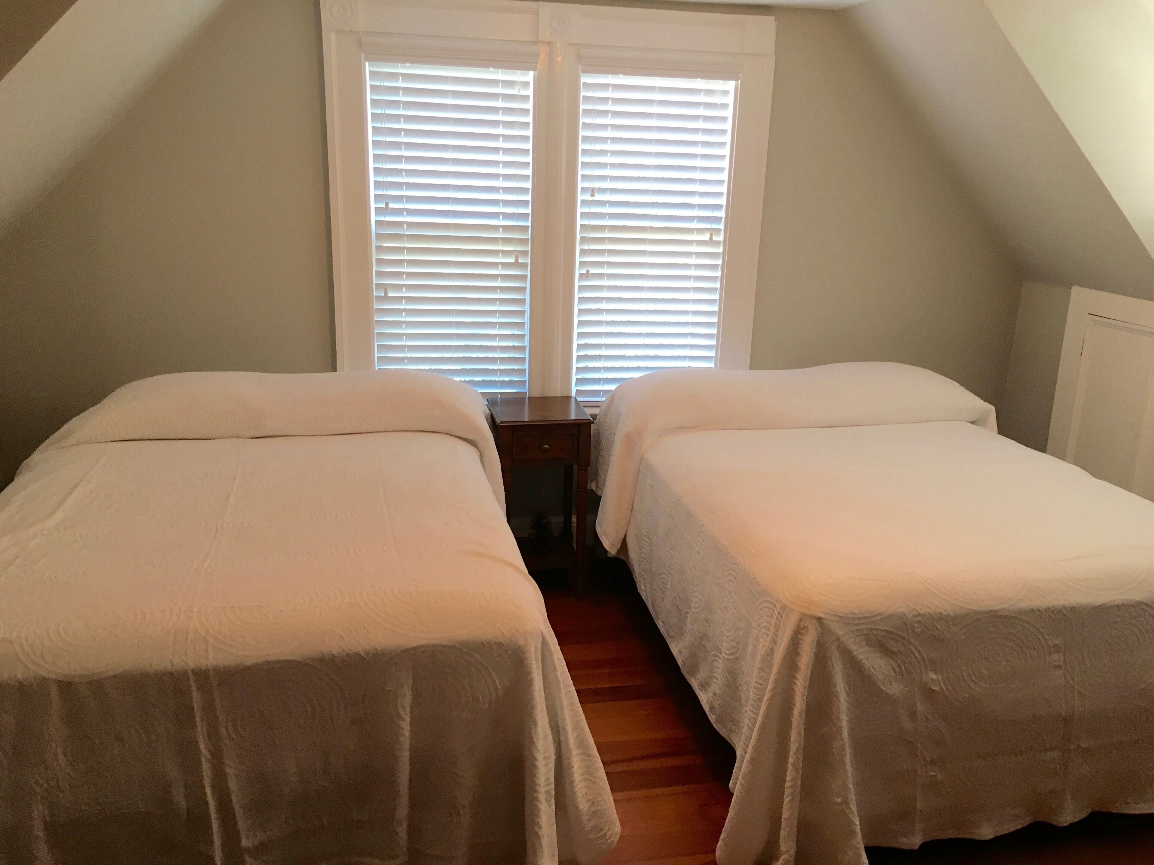 Une chambre avec deux lits, une fenêtre avec des stores et un plancher en bois.