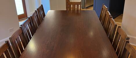 A spacious dining area with a long wooden table and matching chairs, a hanging light fixture, and a kitchen in the background.