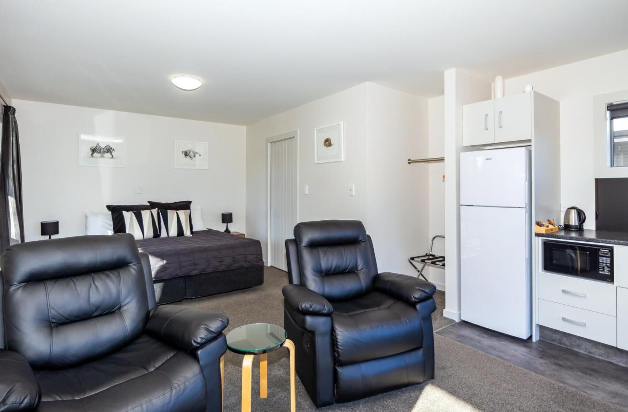 A modern living room with a sofa and two leather armchairs, a small glass table, a bed with a dark-colored duvet, and a kitchenette with a white refrigerator and microwave.