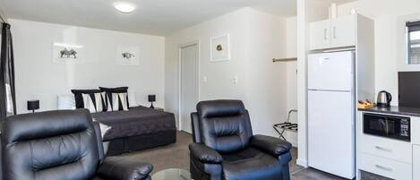 A modern living room with a sofa and two leather armchairs, a small glass table, a bed with a dark-colored duvet, and a kitchenette with a white refrigerator and microwave.