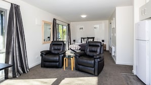 Una sala moderna con dos sofás de cuero negro, una mesa de centro de vidrio y un área de comedor con mesa y sillas. Hay un espejo grande en la pared, un refrigerador y una ventana con cortinas.