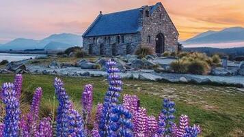 Um edifício de pedra com telhado inclinado, rodeado por um campo de flores roxas.