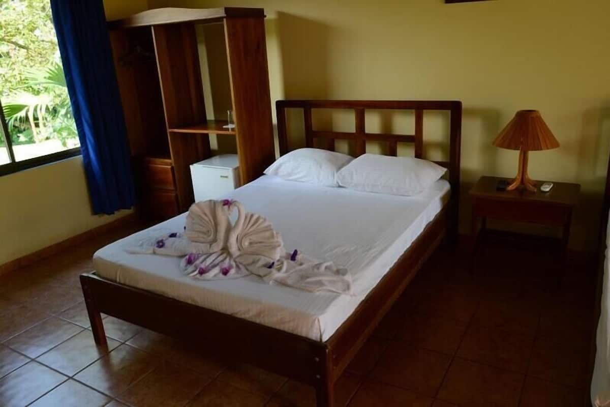 Una cama bien hecha con cabecera de madera, una mesita de noche con lámpara y una pequeña estantería.