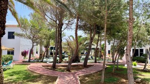 A courtyard with palm trees, a brick pathway, and a building with windows and a balcony. - Aparthotel Playa Parc (Mercadal)