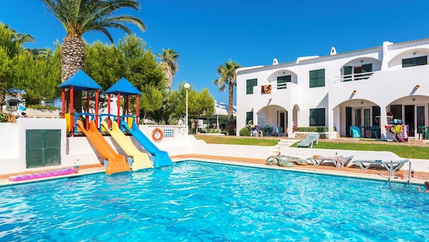 A swimming pool with slides, a palm tree, and a white building with balconies. - Aparthotel Playa Parc (Mercadal)