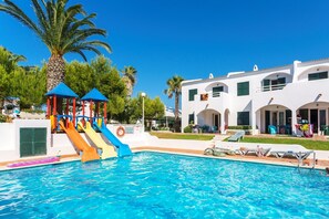 A swimming pool with slides, a palm tree, and a white building with balconies. - Aparthotel Playa Parc (Mercadal)