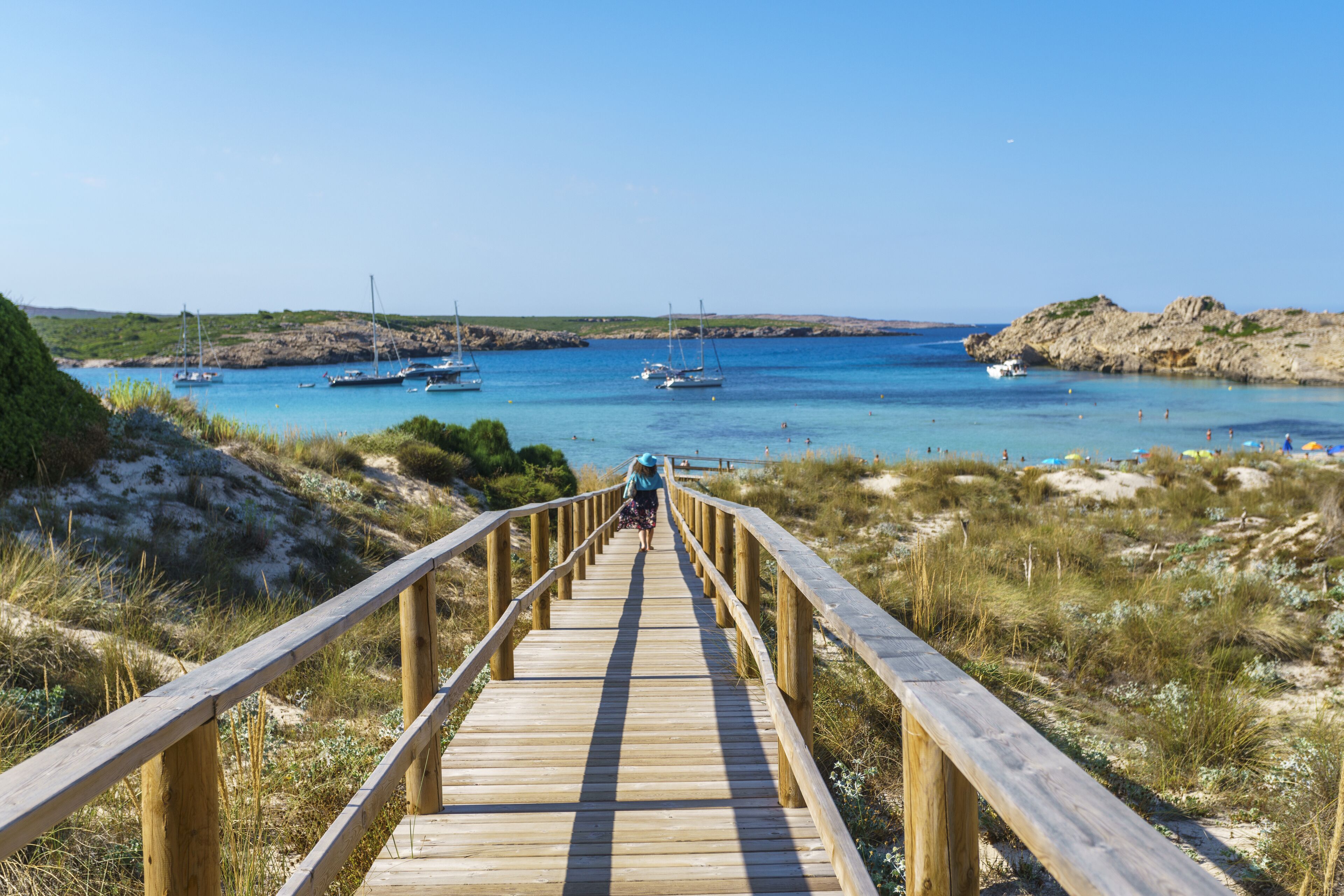 Et træfortov fører til en strand med både og krystalklart, blåt vand.