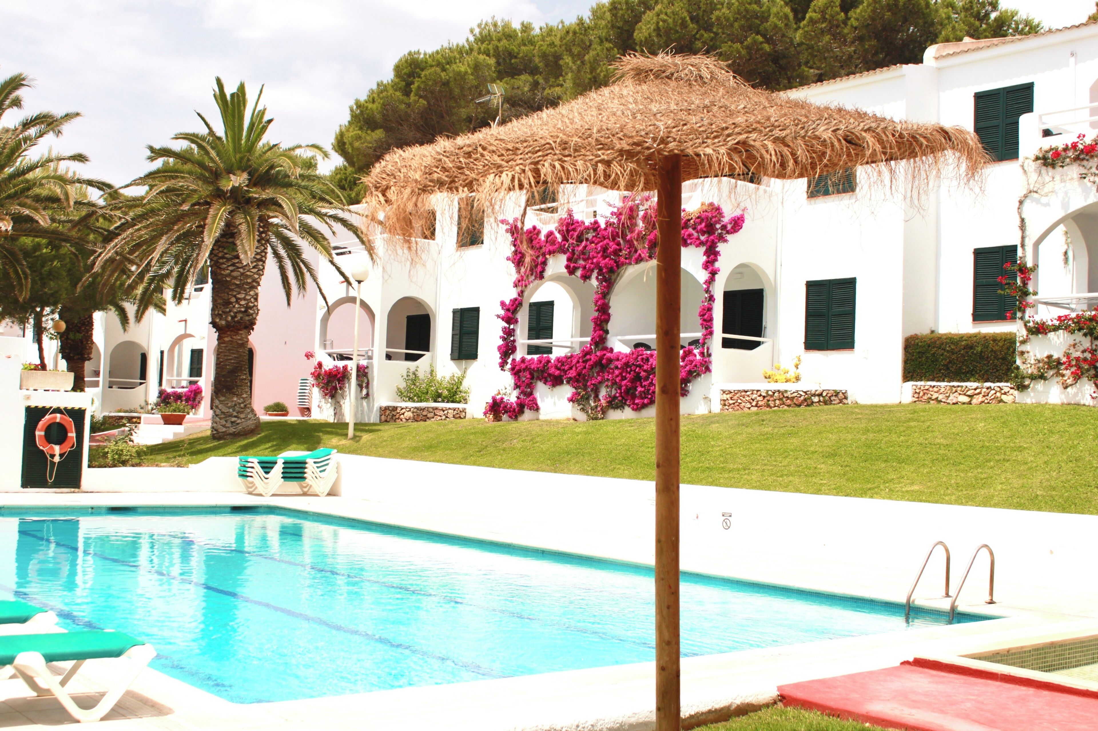 Una piscina con una sombrilla de paja, palmeras y un edificio blanco con contraventanas verdes y flores rosas.