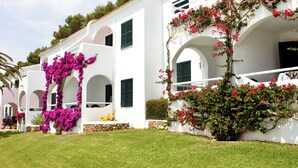 A white building with arched windows and green shutters, surrounded by well-maintained grass and flowering plants. - Aparthotel Playa Parc (Mercadal)