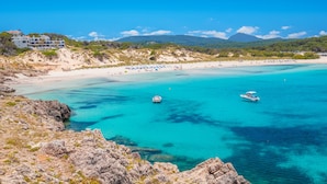 A coastal beach with clear turquoise waters, rocky terrain, and a building with multiple balconies overlooking the beach. - Aparthotel Playa Parc (Mercadal)
